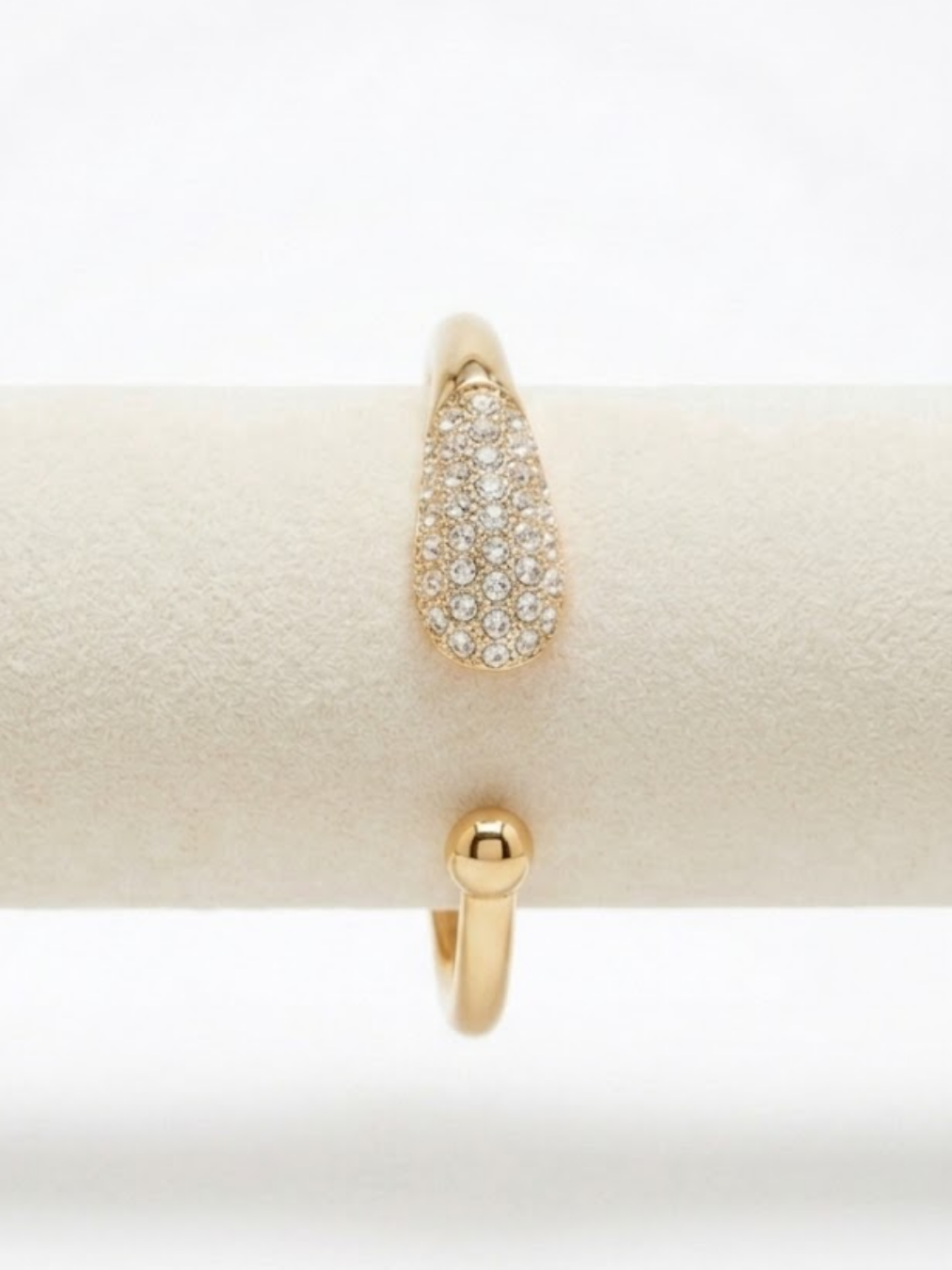Primer plano de brazalete rígido abierto dorado con diseño de gota cubierta de cristales brillantes y detalle de esfera lisa en el extremo opuesto de Angel Salvaje.