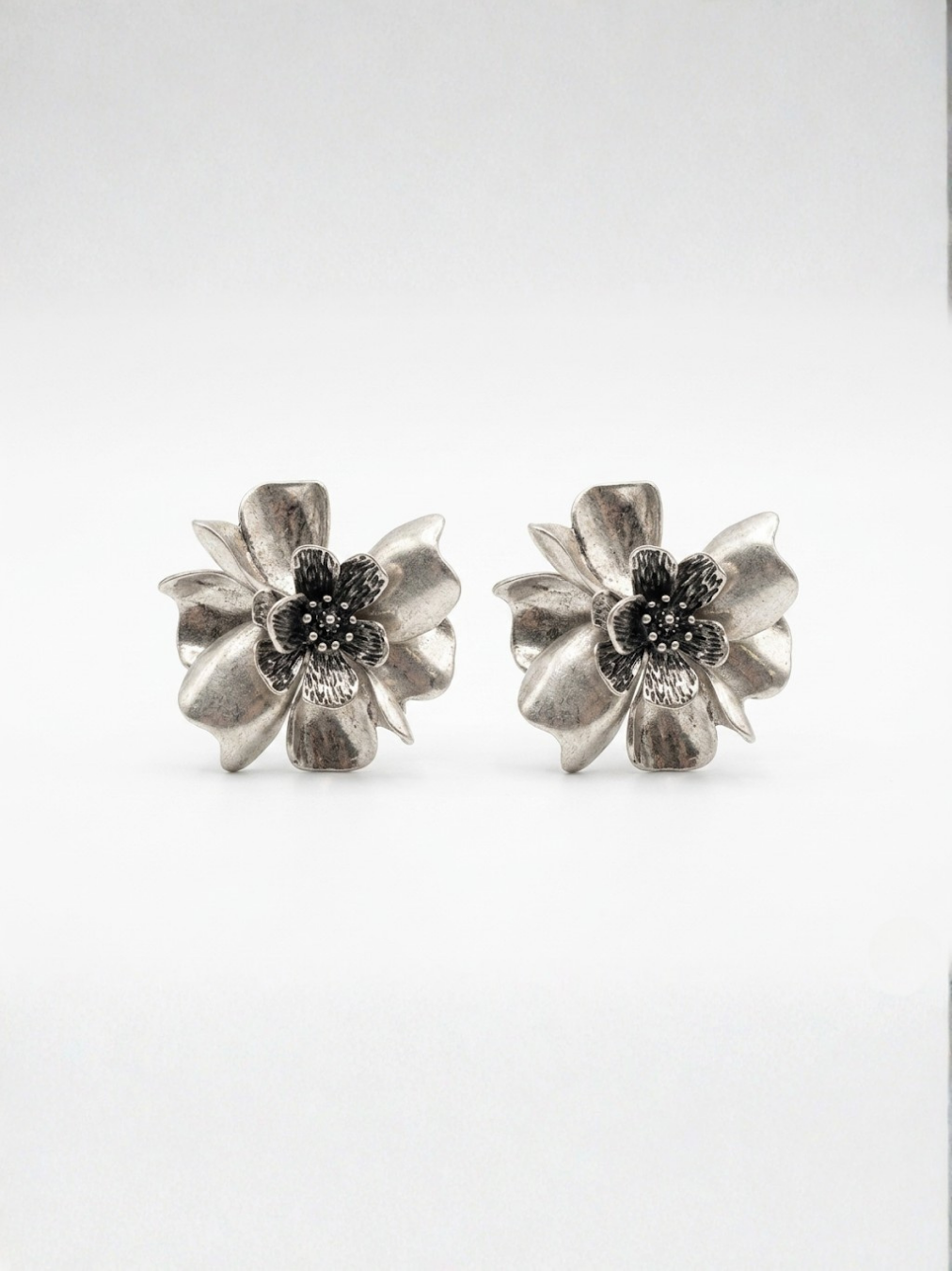Vista frontal de pendientes grandes de botón con diseño de flor tridimensional y pétalos texturizados en tono plateado envejecido de Ángel Salvaje.