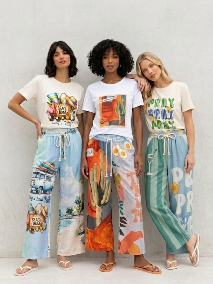 Tres modelos posando juntas con conjuntos casuales de verano compuestos por camisetas blancas con gráficos ilustrados y pantalones anchos fluidos con estampados playeros, de cactus y de rayas en la página de inicio de Ángel Salvaje.