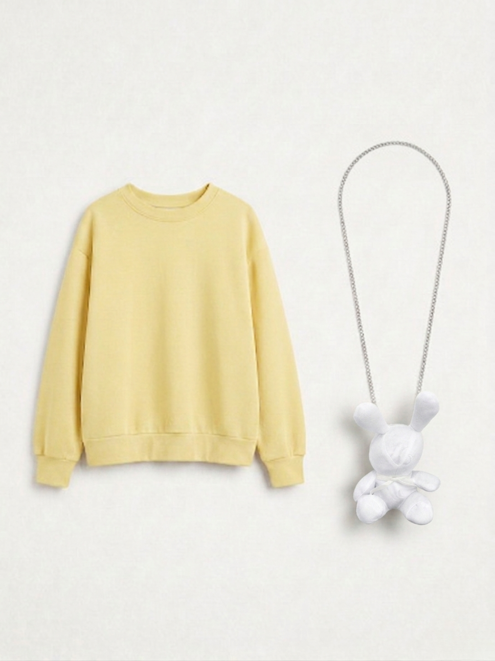 Sudadera amarilla pastel de cuello redondo en plano con un collar de cadena plateada y colgante de conejo de peluche blanco.