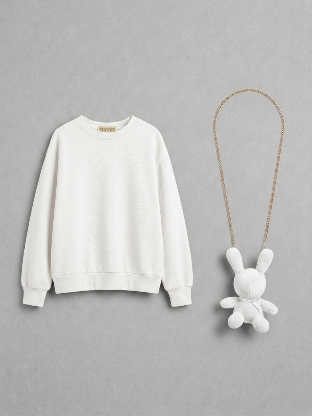 Sudadera oversize de color marfil en plano junto a un collar de cadena dorada con colgante de conejo de peluche blanco.