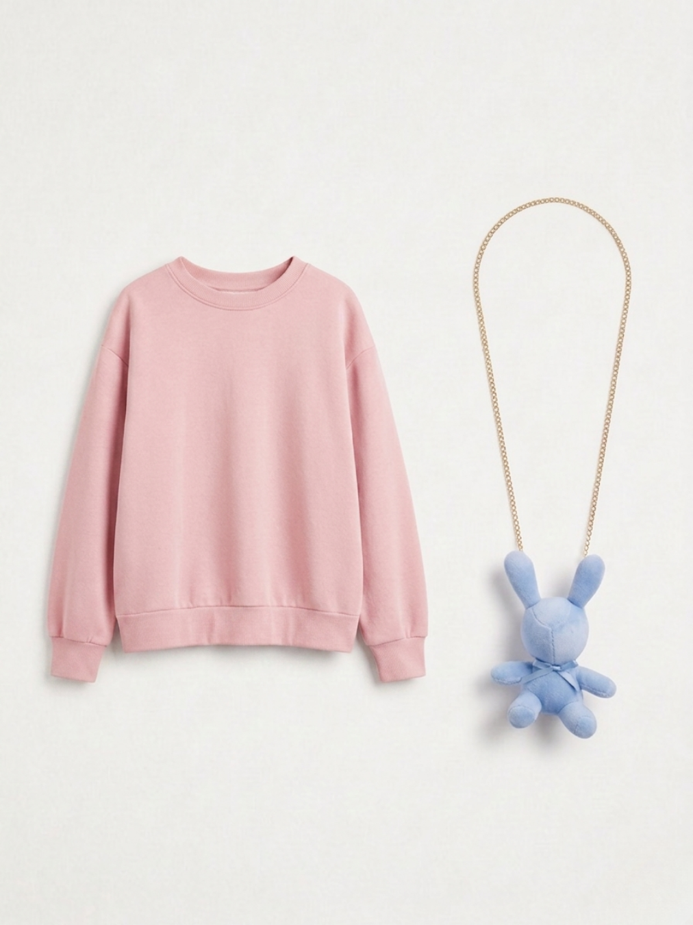 Sudadera oversize rosa claro junto a un accesorio de collar de cadena dorada con un colgante de conejo de peluche azul bebé.