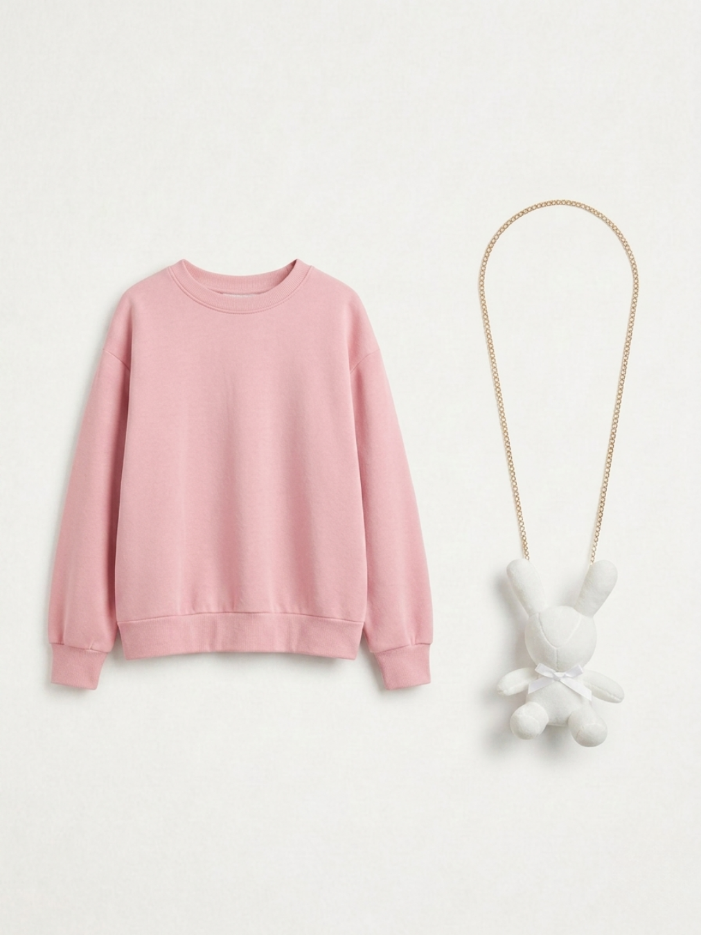 Sudadera rosa pastel de corte relajado presentada en plano con un collar de cadena dorada y colgante de conejo de peluche blanco.